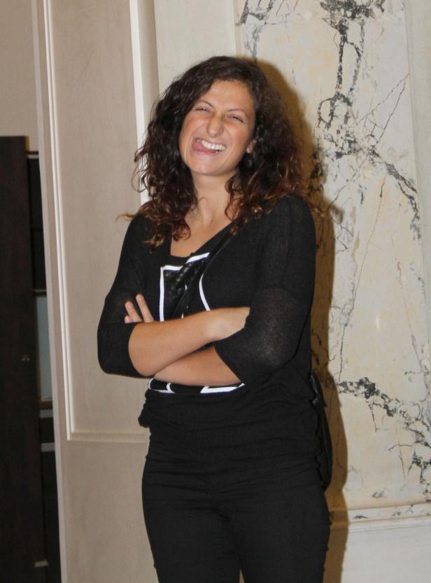 Eine Frau mit lockigem Haar steht mit verschränkten Armen da und streckt die Zunge heraus.