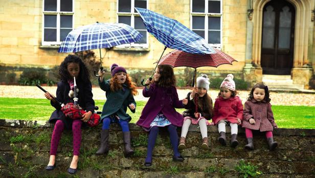 Fünf Mädchen sitzen unter Regenschirmen vor einem Gebäude, eines spielt Dudelsack.