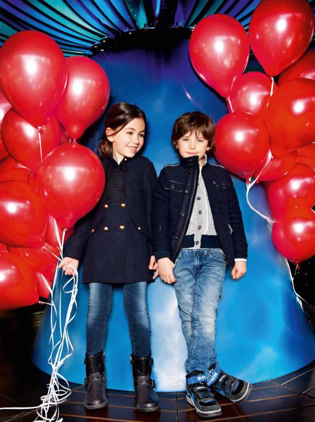 Ein Mädchen und ein Junge stehen vor einem blauen Hintergrund, umgeben von roten Luftballons.