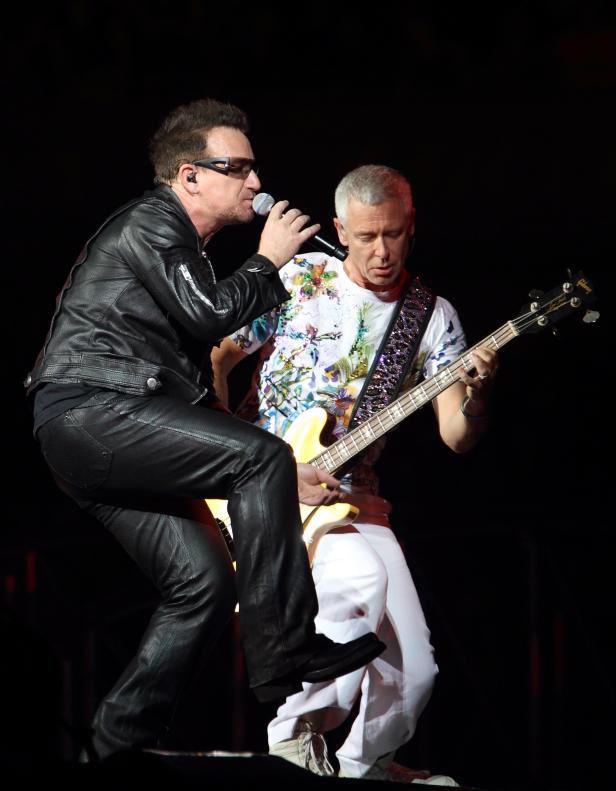 Bono von U2 singt mit Sonnenbrille, während ein Bandmitglied Bass spielt.