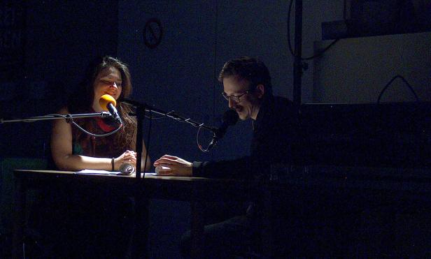 Eine Frau und ein Mann sitzen an einem Tisch mit Mikrofonen und scheinen eine Aufnahme zu machen.