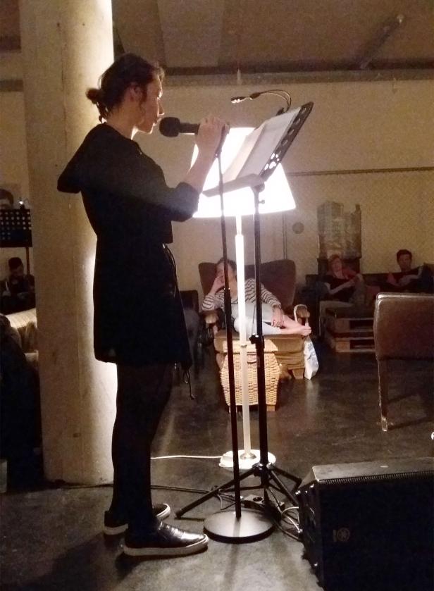 Eine Person liest mit einem Mikrofon vor Publikum von einem beleuchteten Notenständer ab.