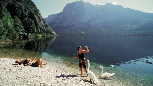Zwei Personen entspannen am Ufer eines Sees, während eine Frau zwei Schwäne füttert. Berge sind im Hintergrund zu sehen.