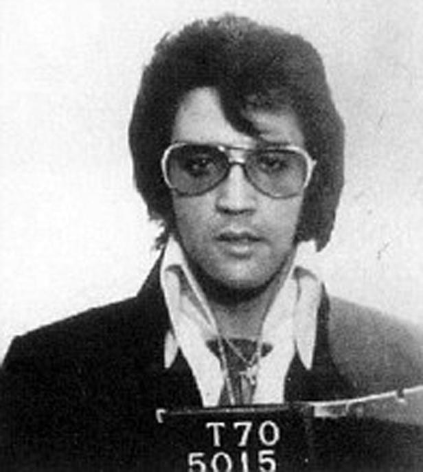 Schwarzweiß-Porträt von Elvis Presley mit Sonnenbrille und einem Schild mit der Aufschrift T70 5015.