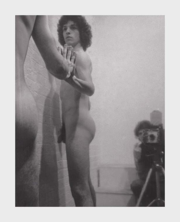 Ein Mann steht nackt vor einem Spiegel, während eine andere Person ihn mit einer Kamera fotografiert.