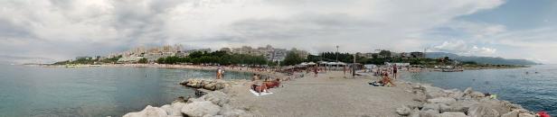 Panoramaaufnahme eines belebten Strandes mit vielen Menschen und Gebäuden im Hintergrund.