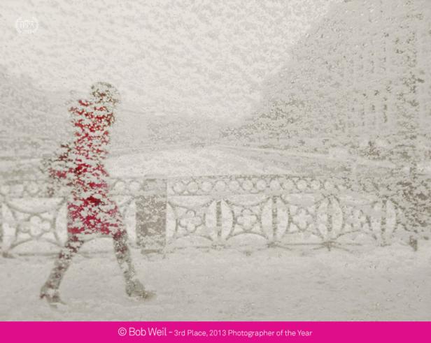 Eine Person in einem roten Mantel geht durch einen Schneesturm an einem Geländer entlang.
