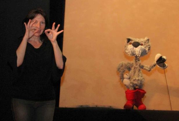 Eine Frau gestikuliert neben einer Katzenpuppe in roten Stiefeln.