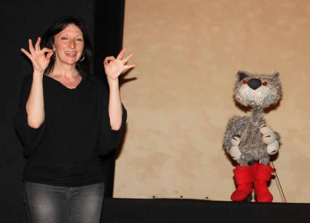 Eine Frau macht Handzeichen neben einer Katzenhandpuppe mit roten Stiefeln.