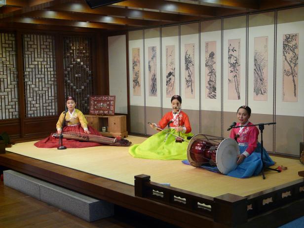 Drei Frauen in traditioneller koreanischer Kleidung spielen Musik auf einer Bühne.