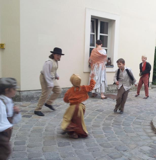 Eine Gruppe von Kindern in historischen Kostümen spielt auf einem gepflasterten Platz.