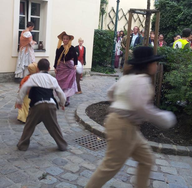 Kinder in historischen Kostümen spielen auf einem gepflasterten Platz.