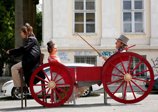 Drei Personen in einem roten, selbstgebauten Wagen auf einer Straße.