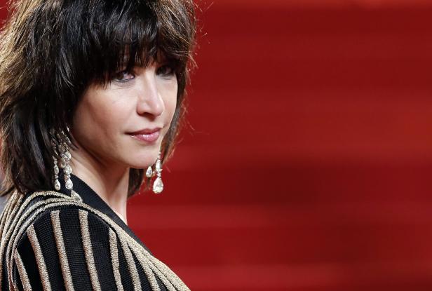 Sophie Marceau mit dunklen Haaren und auffälligen Ohrringen vor rotem Hintergrund.