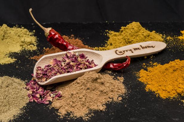 Ein Holzlöffel mit Rosenblättern liegt zwischen verschiedenen Gewürzen und einer roten Chili.