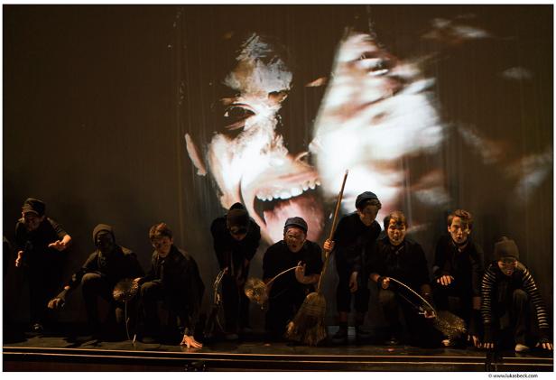 Eine Gruppe von Schauspielern in dunklen Kostümen auf einer Bühne vor einer großen Projektion.