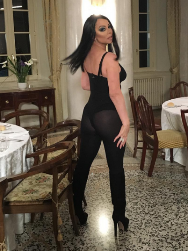 Eine Frau in schwarzem Body und Stiefeln posiert in einem eleganten Raum.