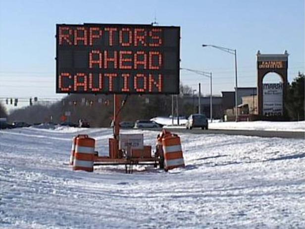 Eine elektronische Anzeigetafel warnt vor „Raptors Ahead Caution“ auf einer verschneiten Straße.