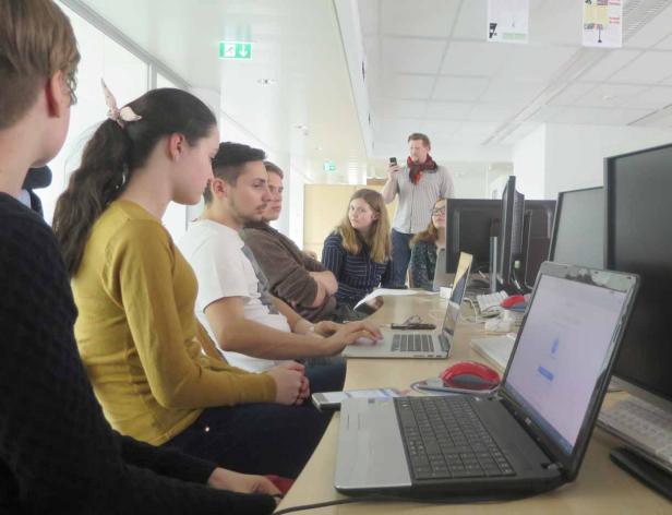 Eine Gruppe Studenten sitzt an Computern, während ein Dozent im Hintergrund steht.