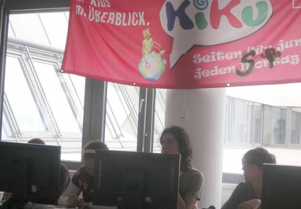 Mehrere Personen sitzen vor Computern unter einem Banner mit der Aufschrift „Kiko“.