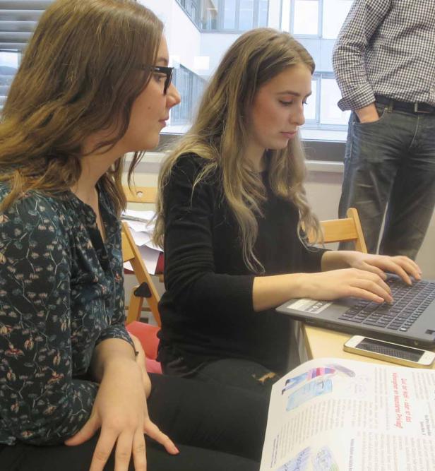 Zwei junge Frauen sitzen an einem Tisch, eine tippt auf einem Laptop.