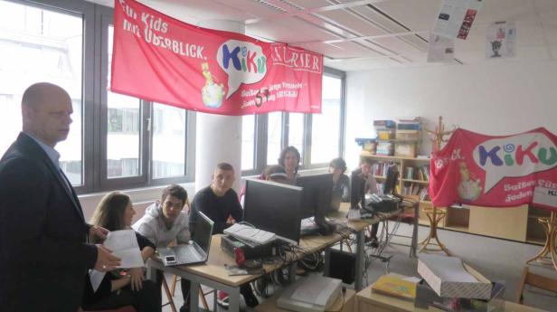 Eine Gruppe von Jugendlichen arbeitet in einem Büro an Computern, im Hintergrund Banner des „KiKu Kurier“.