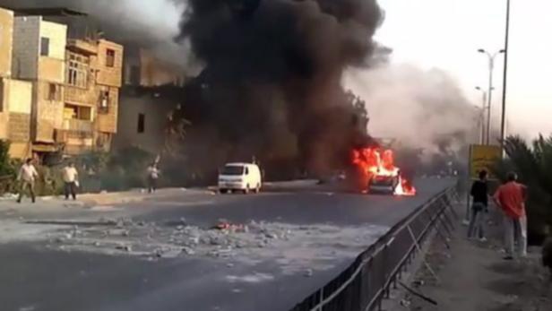 Ein brennendes Auto auf einer Straße, umgeben von Rauch und Trümmern.