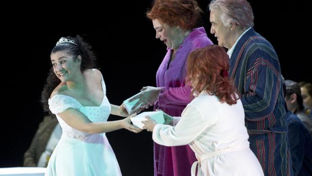 Eine Frau im weißen Kleid und mit Tiara nimmt eine Schachtel von zwei Personen im Bademantel entgegen.