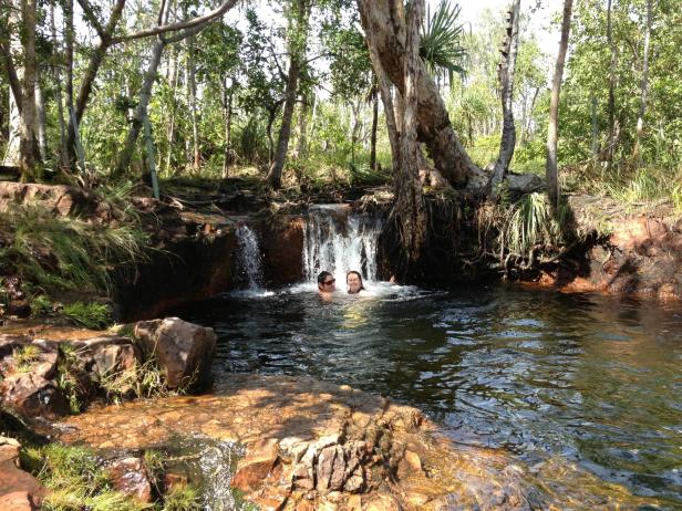 Zwei Personen schwimmen in einem natürlichen Pool unter einem kleinen Wasserfall.