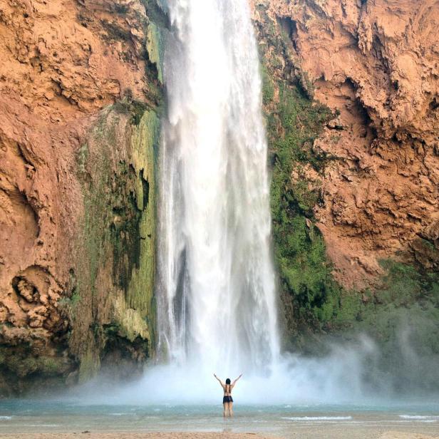 Eine Frau steht mit erhobenen Armen vor einem hohen Wasserfall.