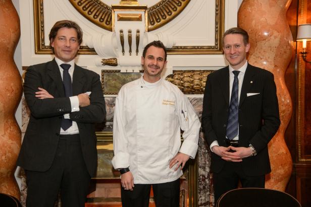 Drei Männer posieren vor einer verzierten Wand, einer davon in Kochuniform.