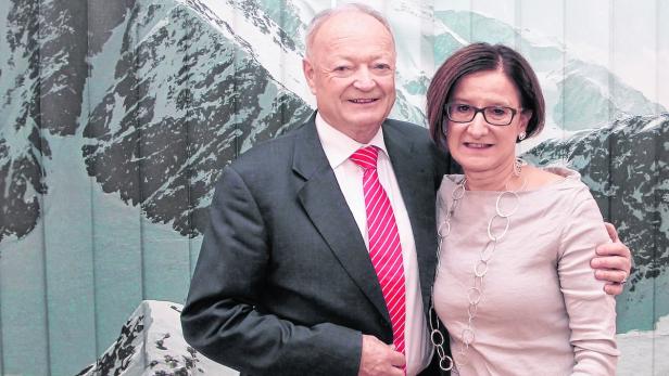 Ein Mann im Anzug und eine Frau mit Halskette posieren vor einem Bergpanorama.