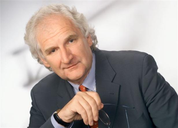 Ein Mann mit grauem Haar und Anzug hält eine Brille in der Hand.