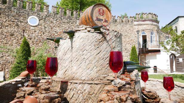 Ein dekorativer Brunnen mit Weinflaschen und großen Weingläsern vor einer Burgmauer.