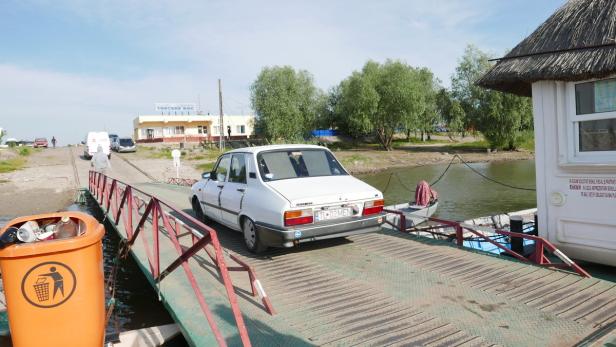 Ein weißer Dacia steht auf einer Fähre, im Hintergrund ein Gebäude mit der Aufschrift „Trecere Bac“.