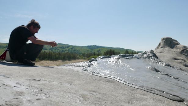 Ein Mann fotografiert einen Schlammvulkan in einer hügeligen Landschaft.