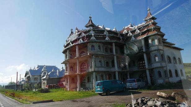 Ein großes, noch unfertiges Gebäude mit aufwendiger Dachkonstruktion und mehreren geparkten Autos davor.