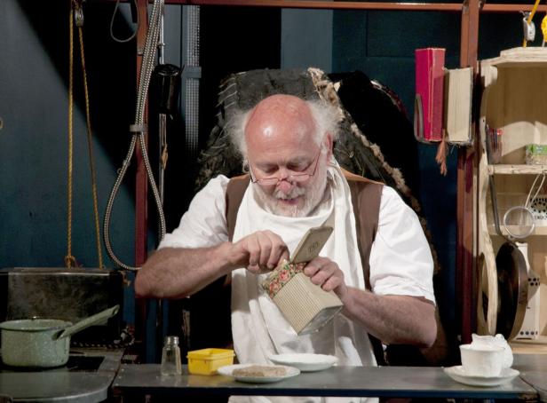 Ein älterer Mann mit Brille öffnet eine verzierte Dose in einer Küche.