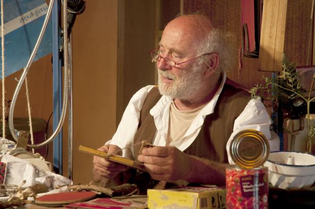 Ein älterer Mann mit Brille fädelt eine Schnur durch ein Holzstück.