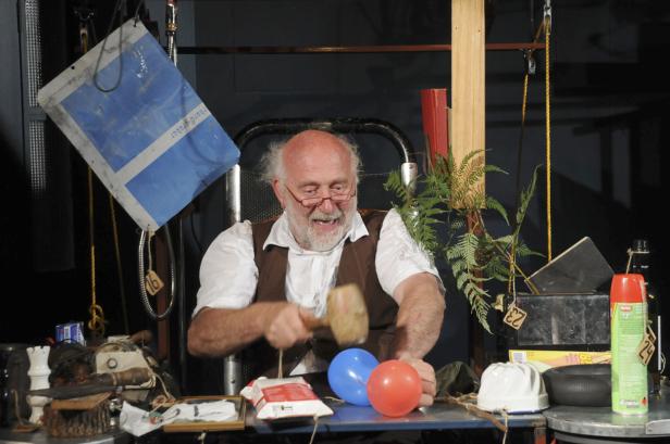 Ein Mann mit Brille und Weste schlägt mit einem Hammer auf zwei Ballons.