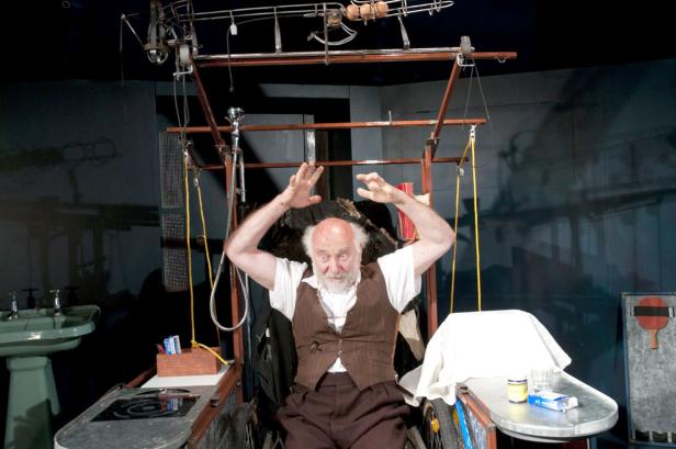 Ein Mann mit erhobenen Händen sitzt in einer Apparatur, die an eine verrückte Erfindung erinnert.