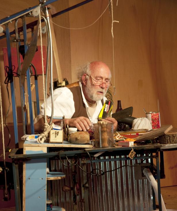 Ein älterer Mann mit Brille arbeitet an einem Tisch voller Werkzeuge und Materialien.