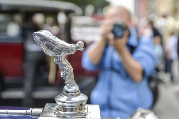 Die Kühlerfigur „Spirit of Ecstasy“ eines Rolls-Royce im Fokus, während eine Person im Hintergrund ein Foto macht.