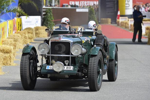 Ein grüner Oldtimer mit zwei Personen fährt auf einer asphaltierten Straße.