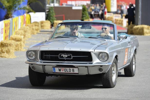 Ein silberner Ford Mustang Oldtimer mit zwei Insassen fährt auf einer Straße.