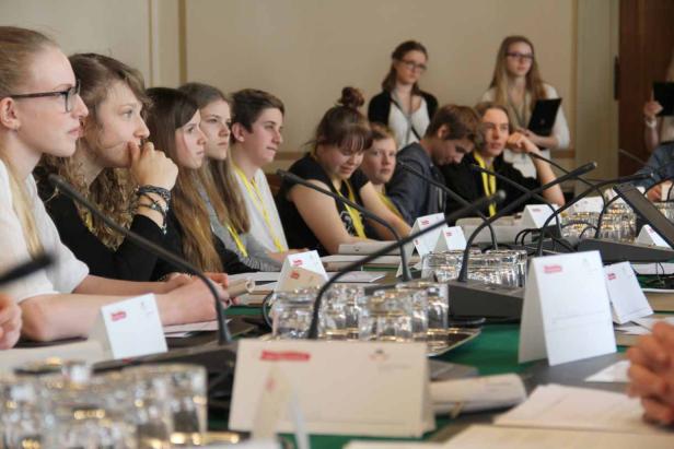 Eine Gruppe junger Leute sitzt an einem Tisch mit Mikrofonen, möglicherweise bei einer Konferenz.