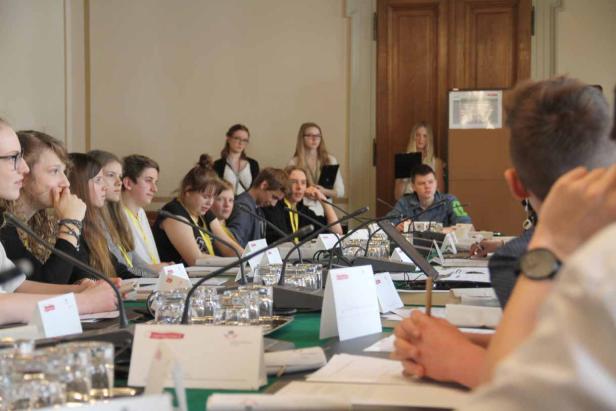 Eine Gruppe junger Leute sitzt an einem Tisch mit Mikrofonen bei einer Konferenz.