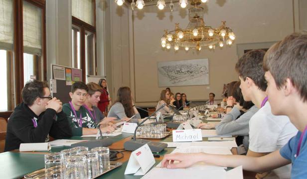 Jugendliche sitzen an einem Tisch mit Mikrofonen in einem Konferenzraum.