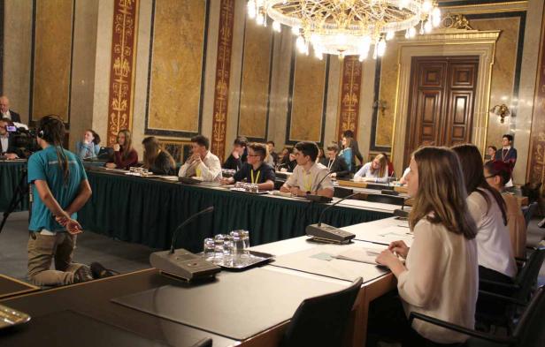 Jugendliche sitzen an Tischen in einem prunkvollen Saal, während ein Kameramann kniet und filmt.