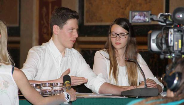 Ein junger Mann und eine junge Frau sitzen an einem Tisch mit Mikrofonen vor einer Kamera.